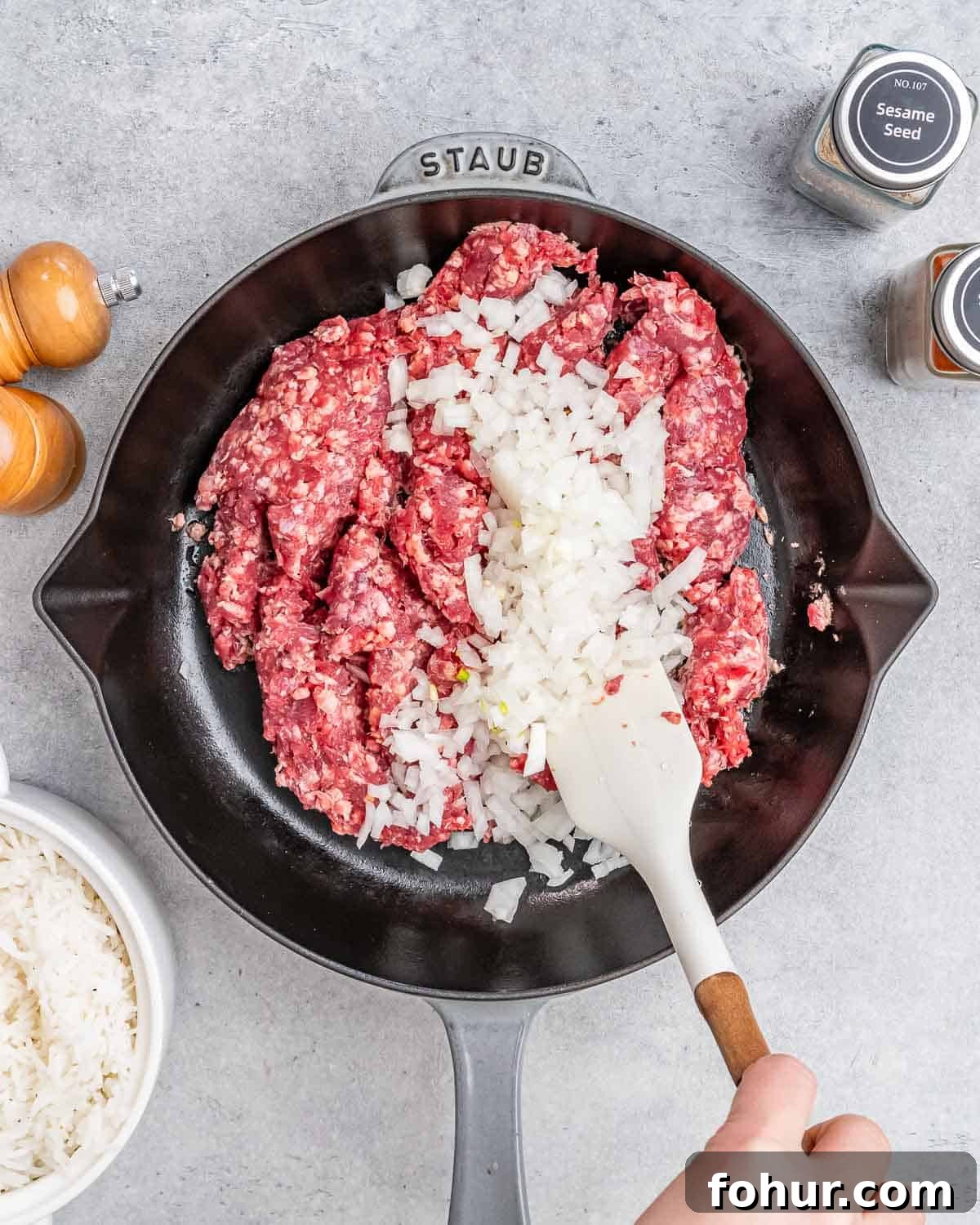 A white spatula stirring onions and raw ground beef in a skillet.