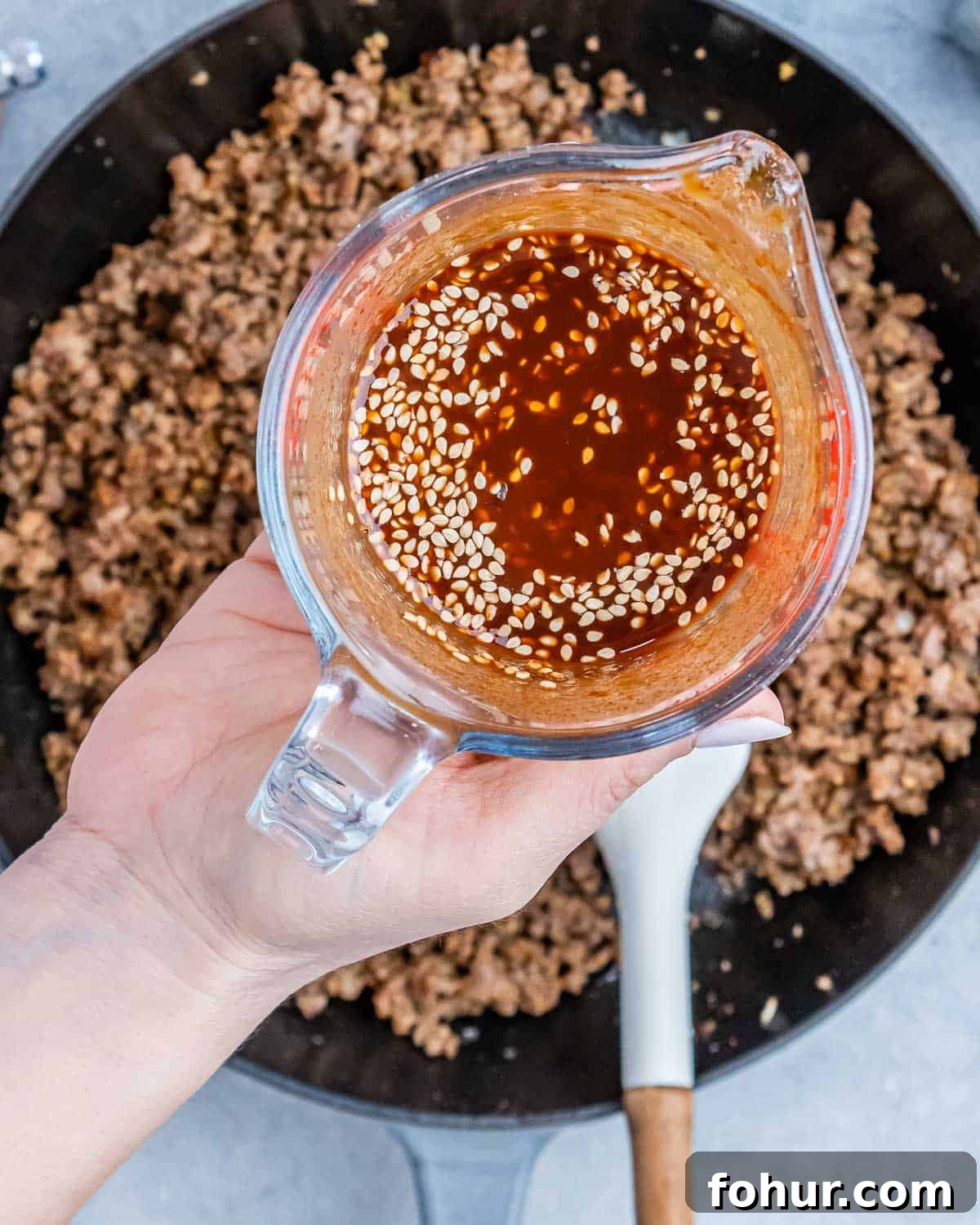 A hand holding liquid over a skillet of ground beef.