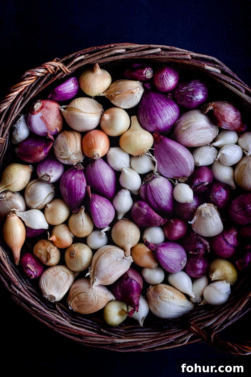 Shallot Confit Recipe: Jammy Slow-Roasted Condiment for Any Dish 3 Basket filled with raw shallots, garlic and tiny onions, in shades of purple, gold and white on a dark background.